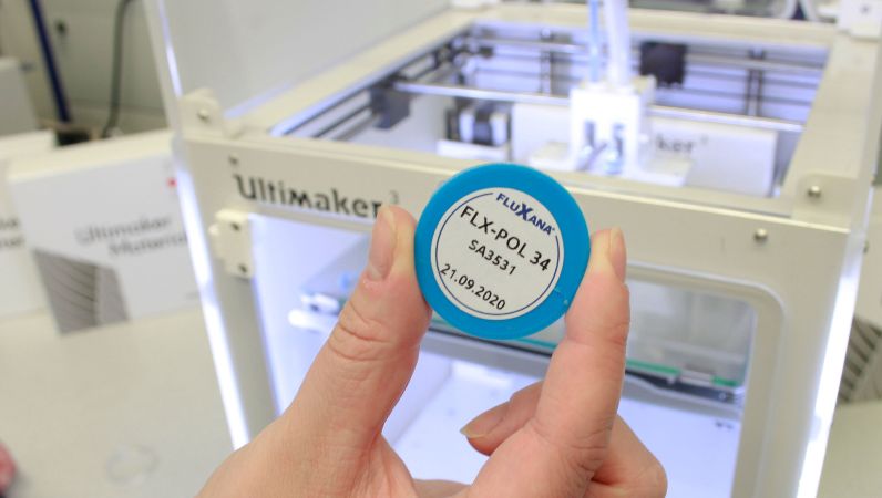 Ein türkiser Probenkörper aus der Materialbibliothek; im Hintergrund der 3-D-Drucker.